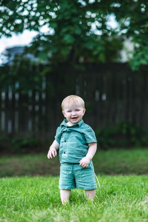 Smiling toddler boy in teal outfit standing on green grass in a backyard.