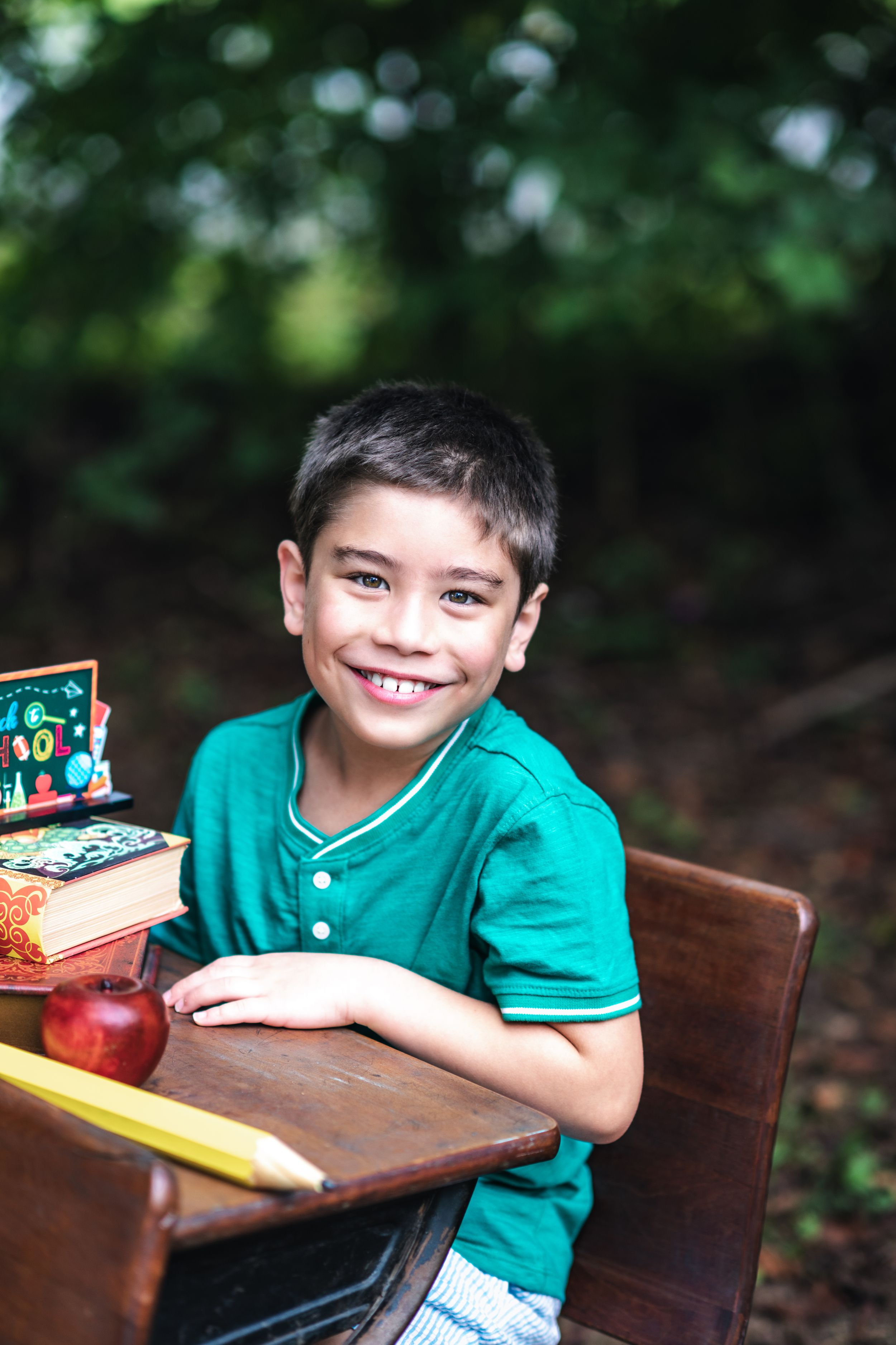 Student sitting at wooden desk with school supplies smiling in green shirt outdoors.