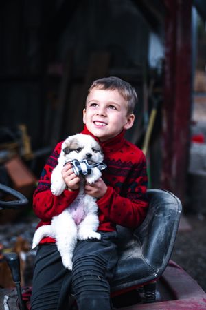 Young boy in red sweater holding small white puppy while sitting on leather seat.