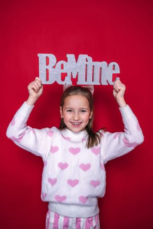 Young girl in heart sweater holds up 'Be Mine' text against red background.
