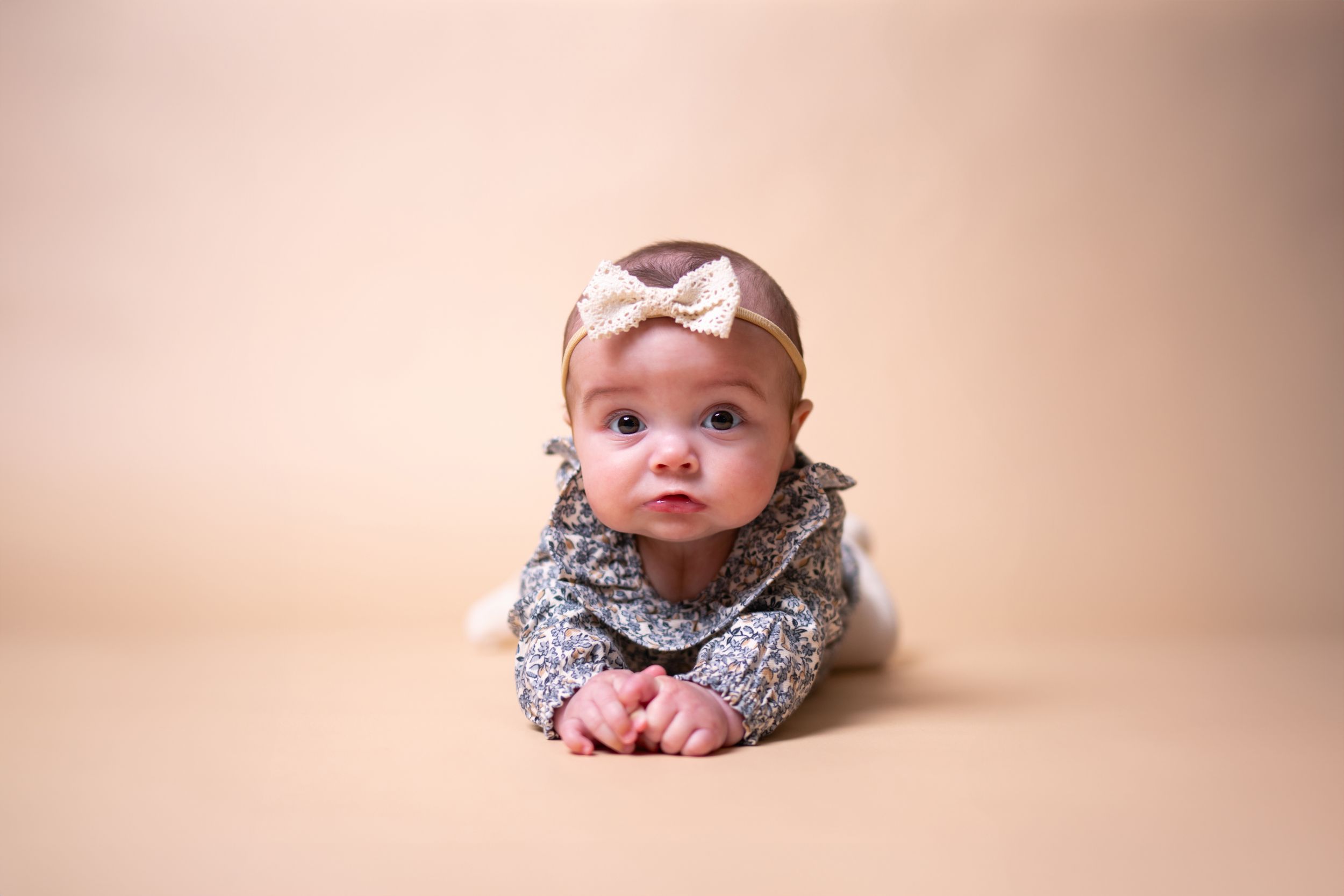 Baby wearing floral dress and bow headband lying on beige backdrop during professional photoshoot.