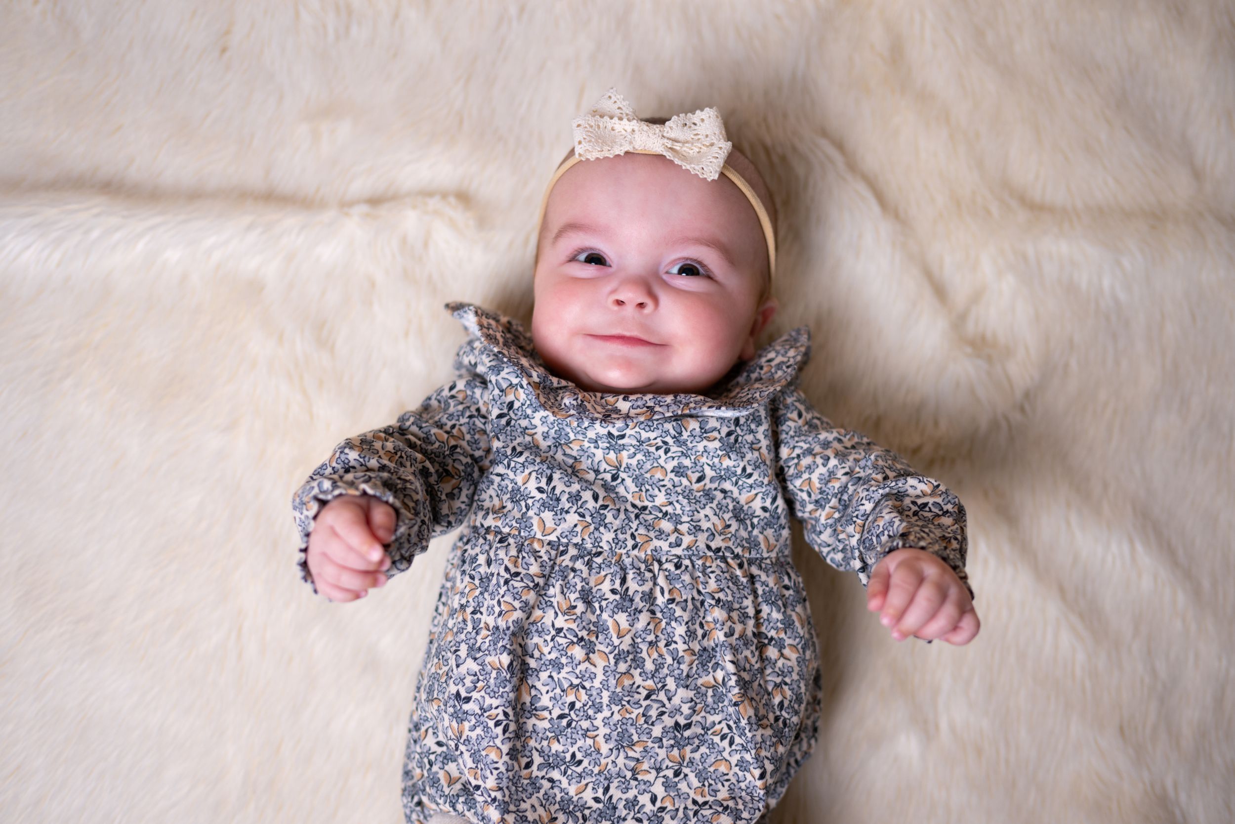 Infant in floral dress and pink bow lying on cream colored blanket.