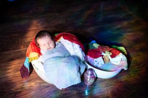 Sleeping newborn wrapped in white blanket with rainbow decorations on wooden floor.