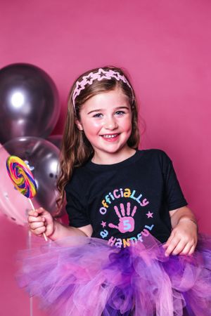 Girl in tutu and birthday shirt poses with lollipop against pink background with balloons.