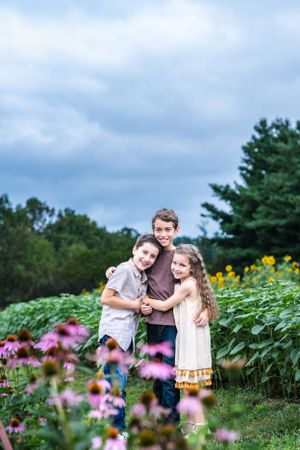 Three children hugging in garden with purple coneflowers and sunflowers.