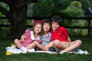 Three happy children sitting together on grass under a tree, smiling and hugging outdoors.