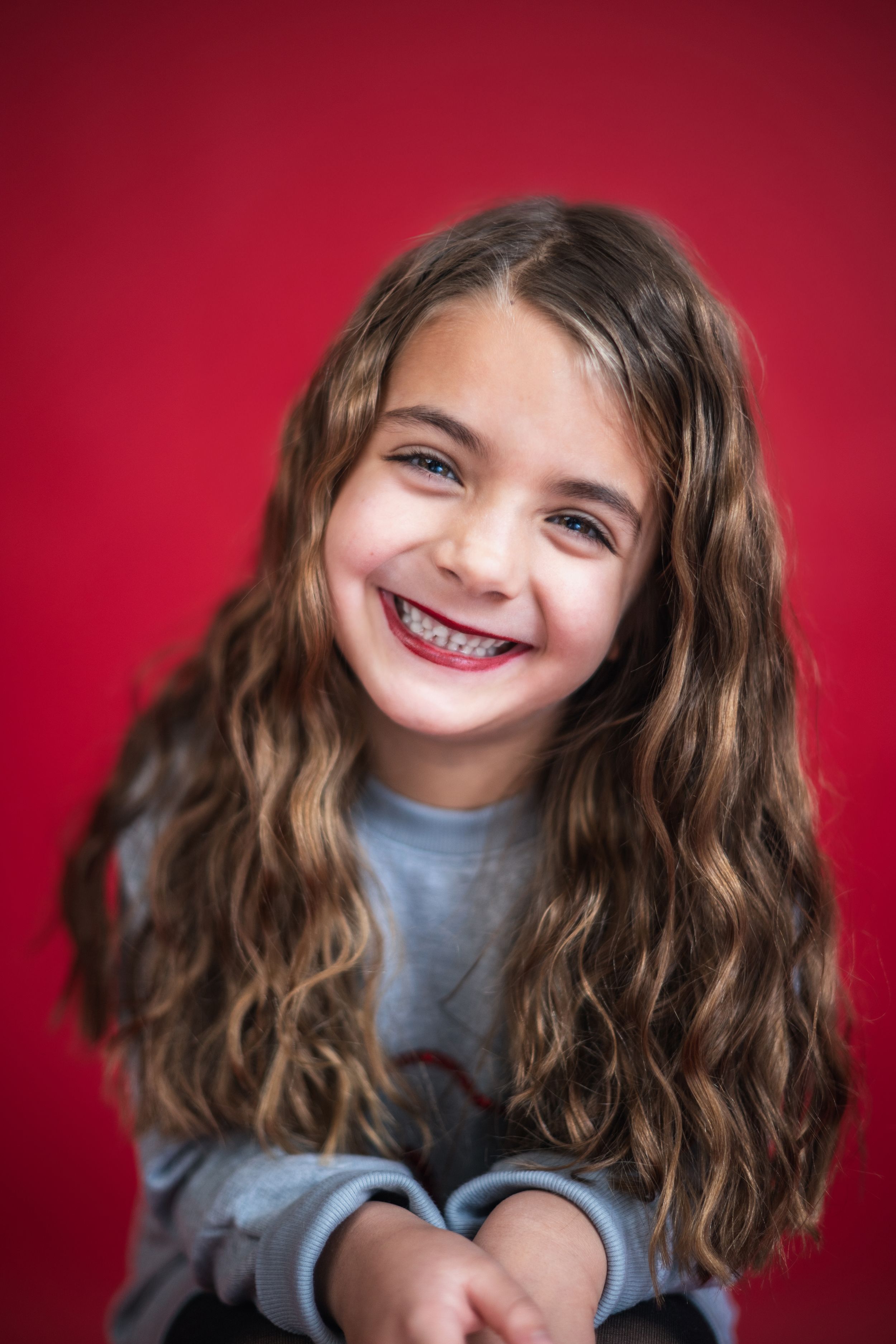 Smiling young girl with long curly brown hair and braces posing against a vibrant red background.