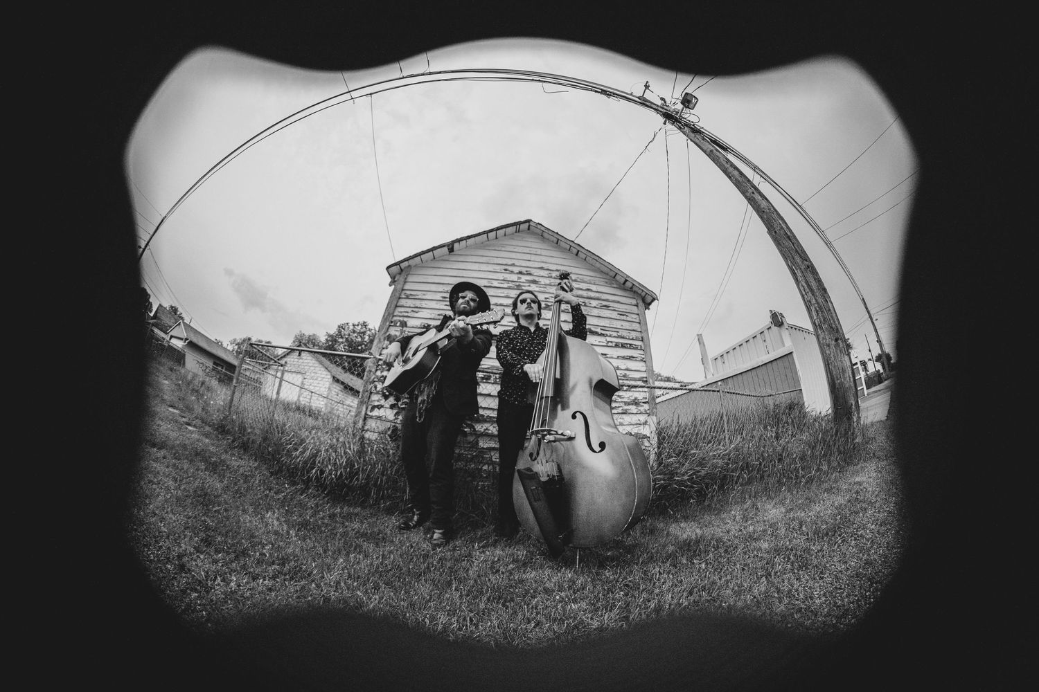 Black and white fisheye photo shows a couple walking past a rustic house carrying vintage luggage in a pastoral setting.