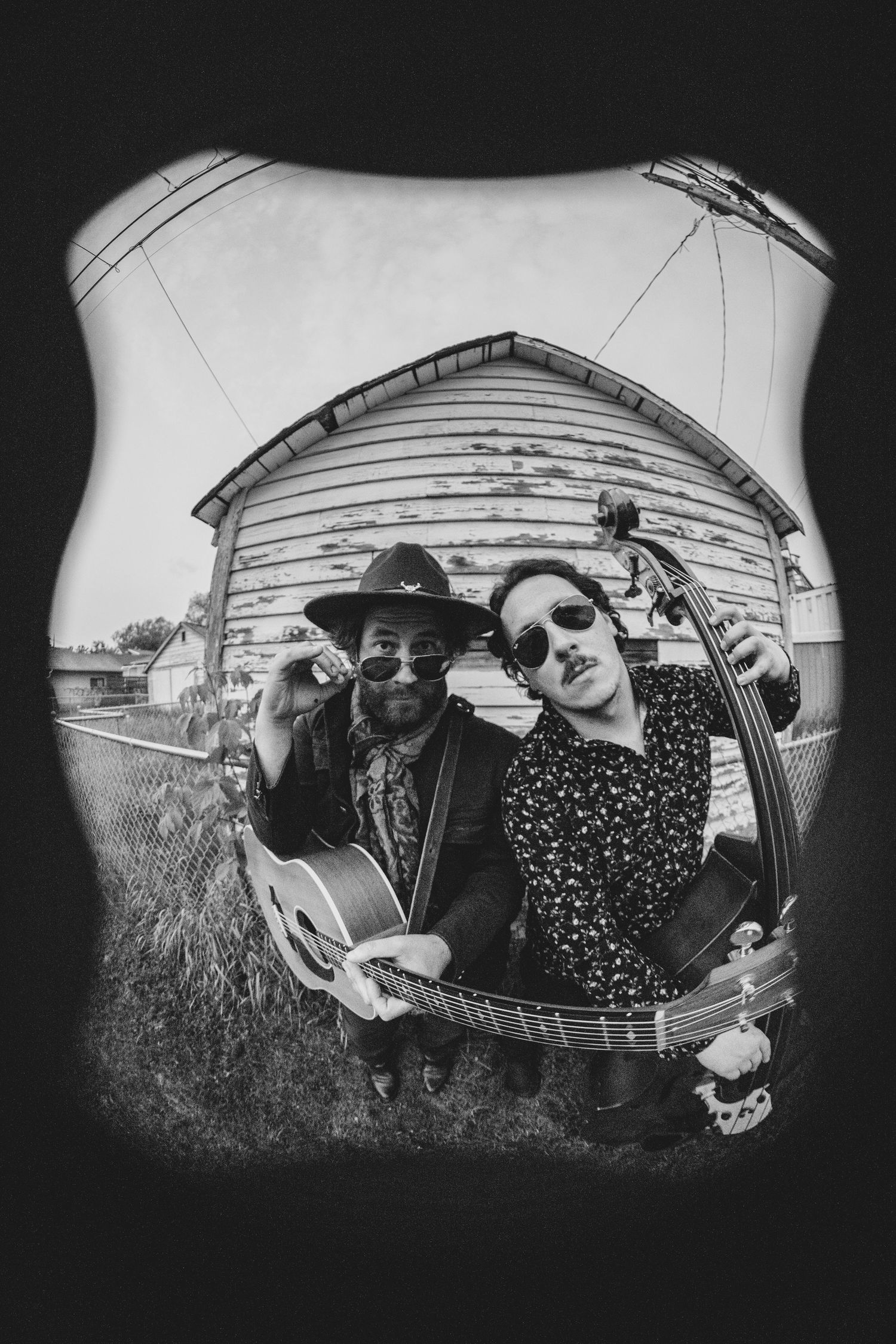 Black and white fisheye photo of two musicians with a guitar posing together in front of a rustic wooden building.