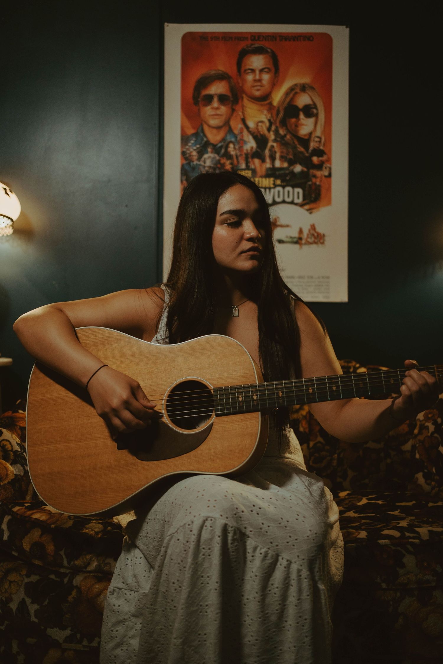 Young woman in white dress playing acoustic guitar on a couch with a Once Upon a Time in Hollywood poster behind her.