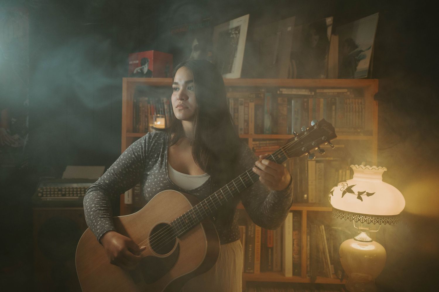 Young woman playing acoustic guitar in a moody, dimly lit room with bookshelves and a vintage oil lamp.