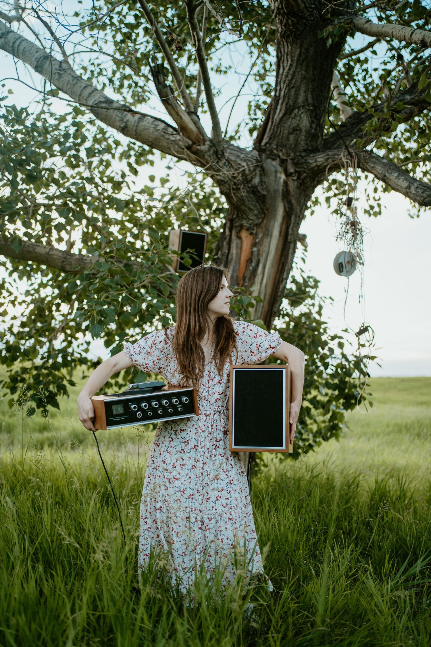 Person in floral dress holding chalkboards while standing by a large tree in a green meadow.