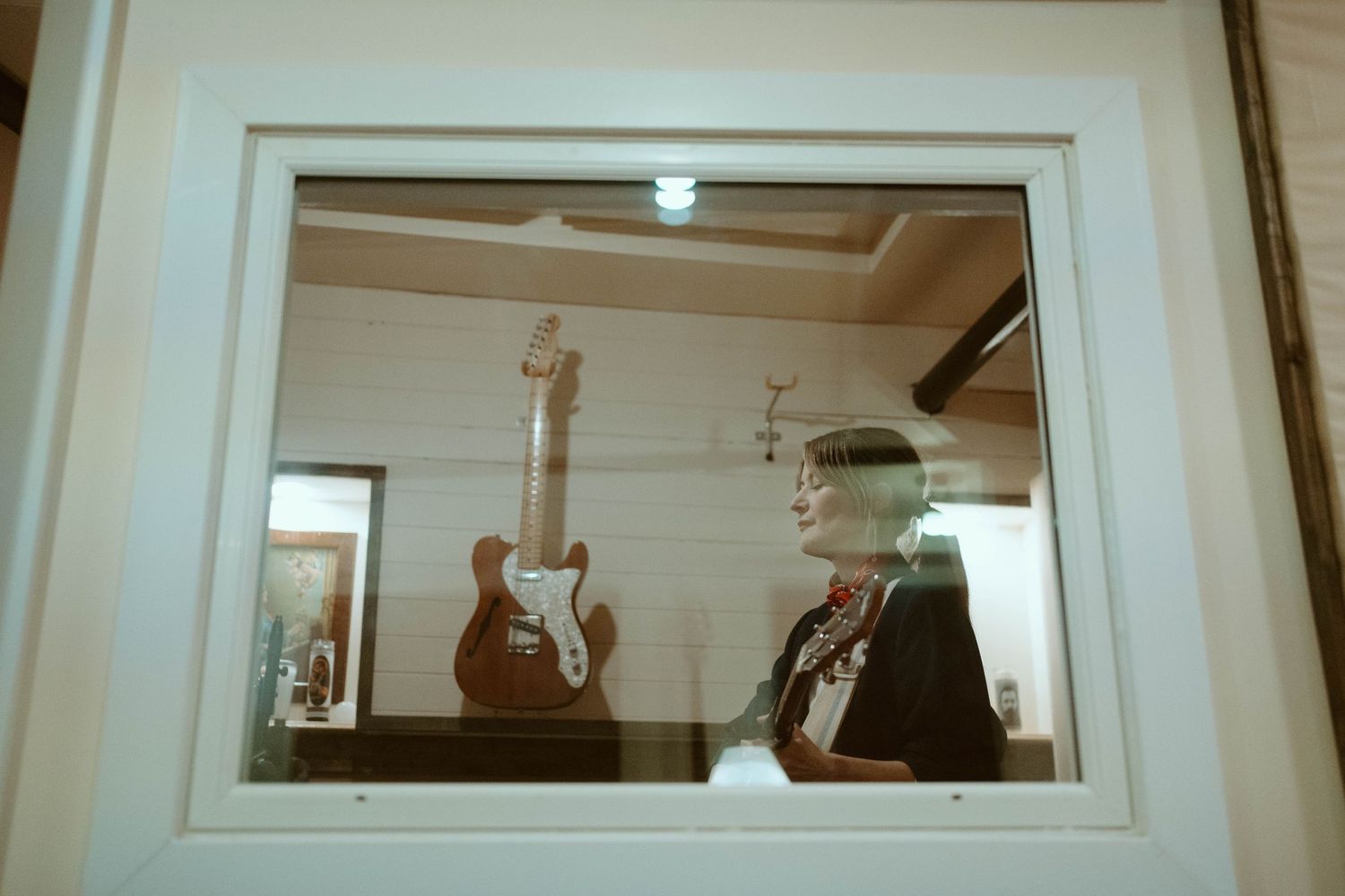 Person reflected in glass window beside a vintage brown electric guitar displayed on a white wall.