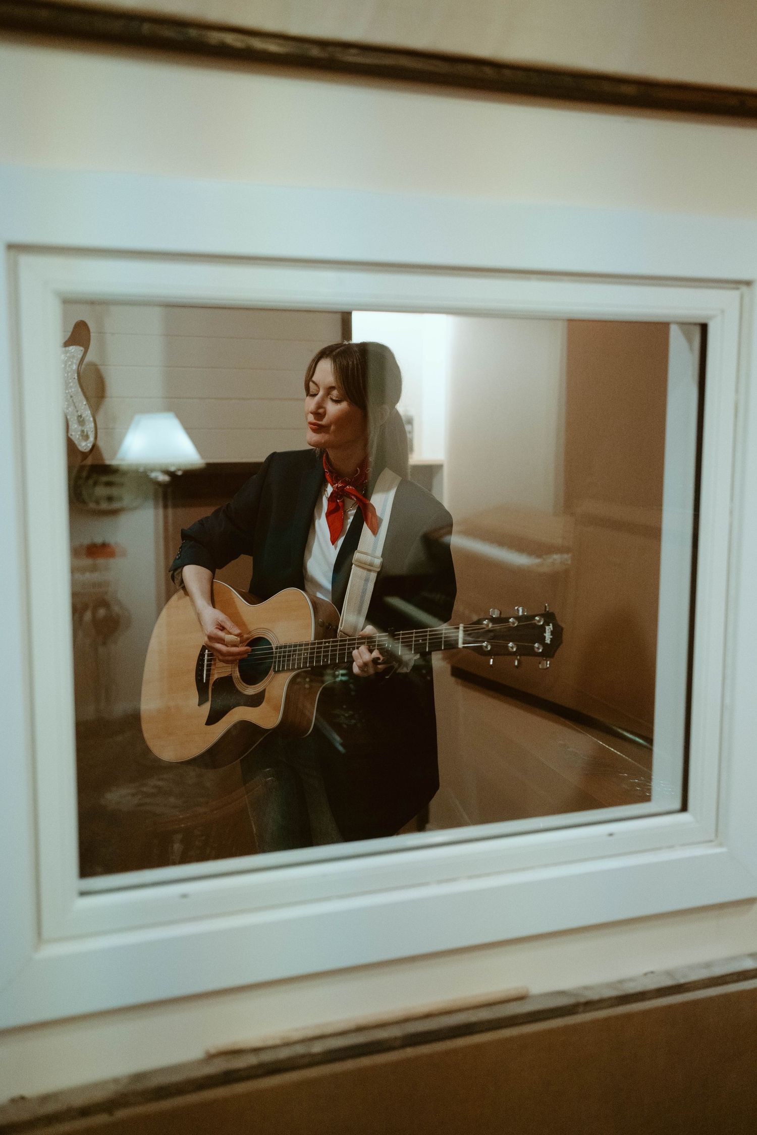 A woman in a black blazer and red scarf plays an acoustic guitar, viewed through a window reflection indoors.