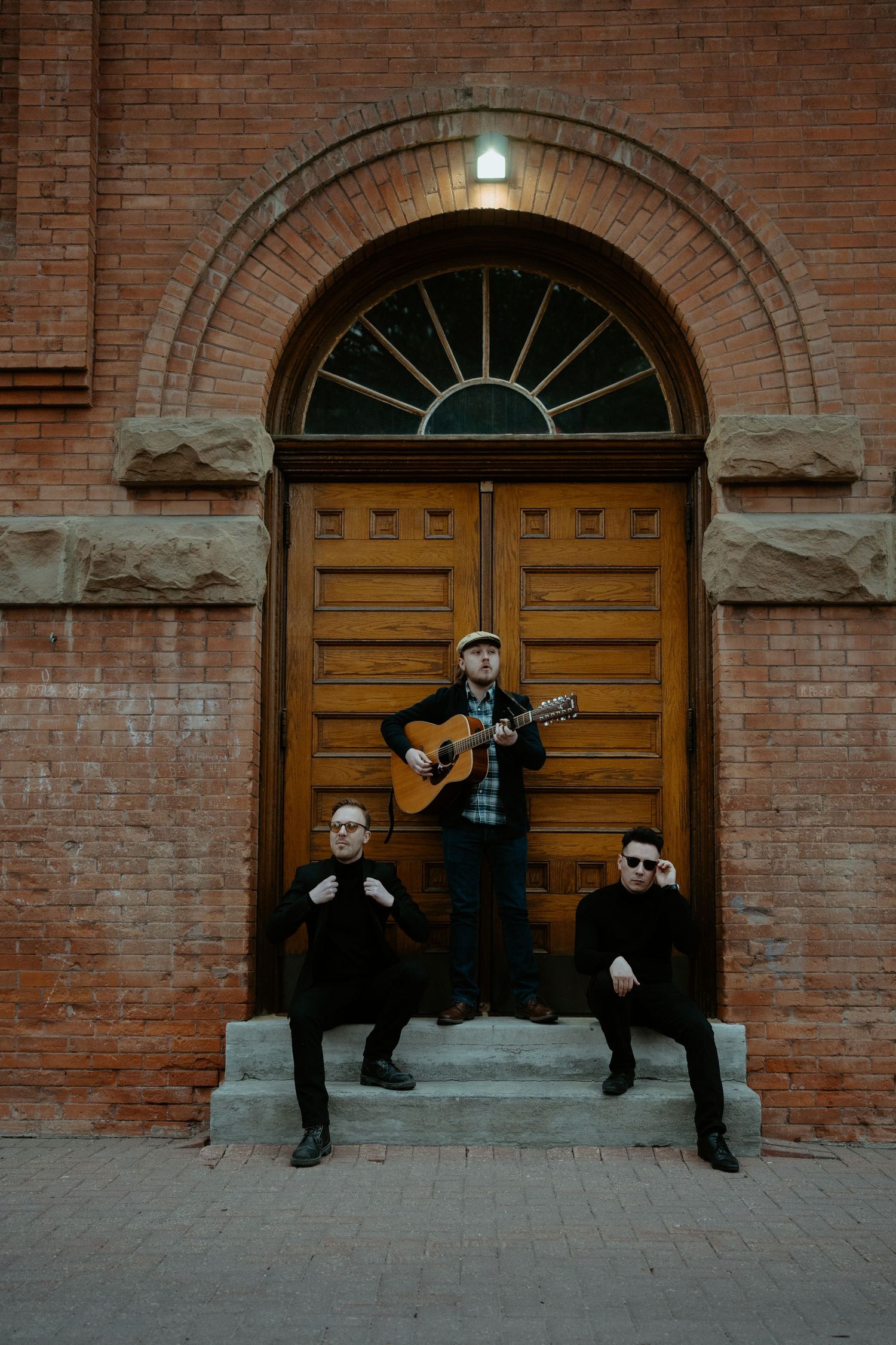 Three musicians pose with a guitar in front of an ornate brick doorway.