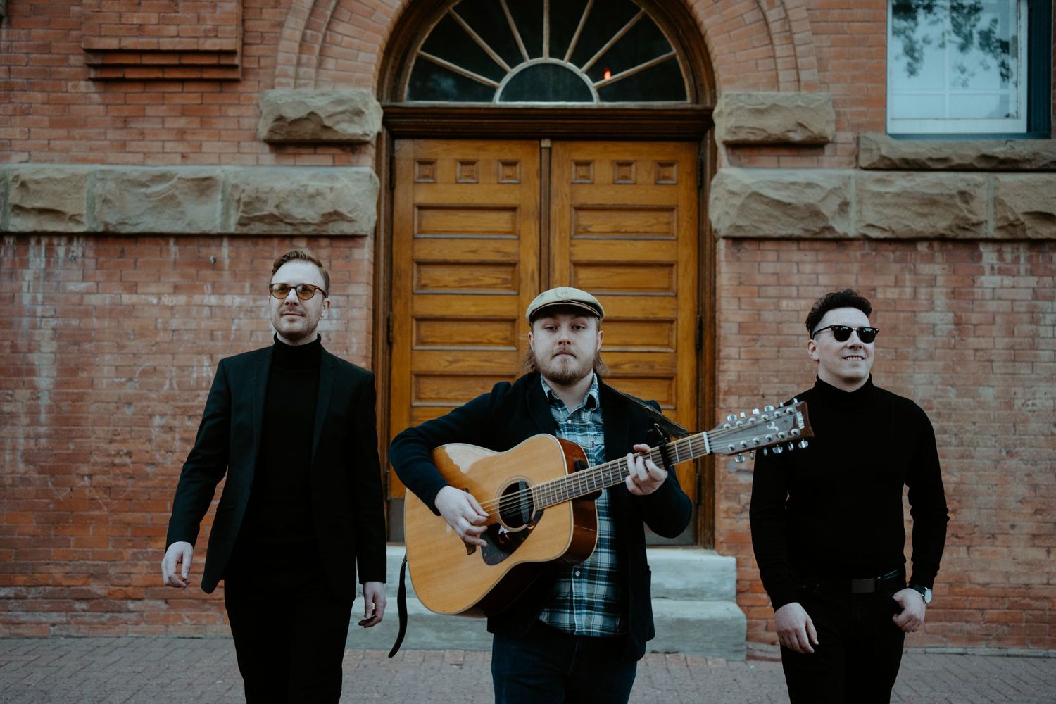 Three musicians in black attire pose with acoustic guitar in front of historic brick building.