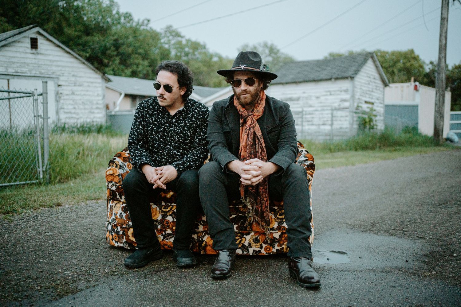 Two musicians in dark attire and western wear sit together on wooden benches in front of white buildings on a rainy day.