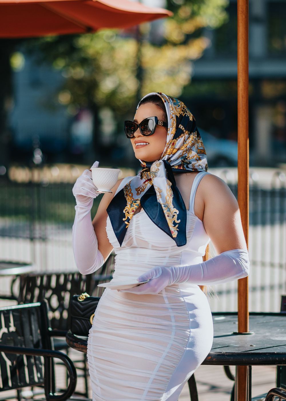 Elegant woman in white dress and patterned headscarf enjoys coffee at an outdoor cafe terrace on a sunny day.