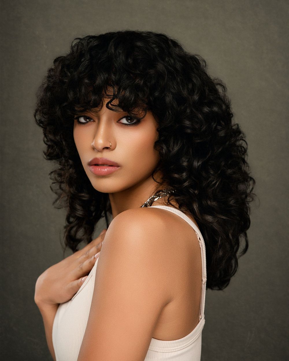 Fashion portrait of a model with voluminous dark curly hair wearing a white tank top against a gray studio backdrop.