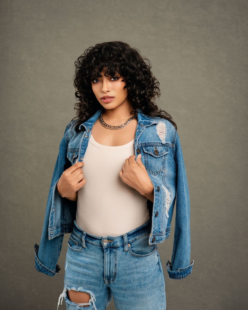 Model in distressed denim jacket and ripped jeans poses against gray backdrop in casual fashion portrait.