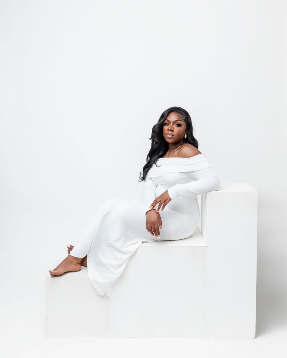 Elegant fashion portrait on white steps featuring off-shoulder white jumpsuit and graceful pose against minimal background.