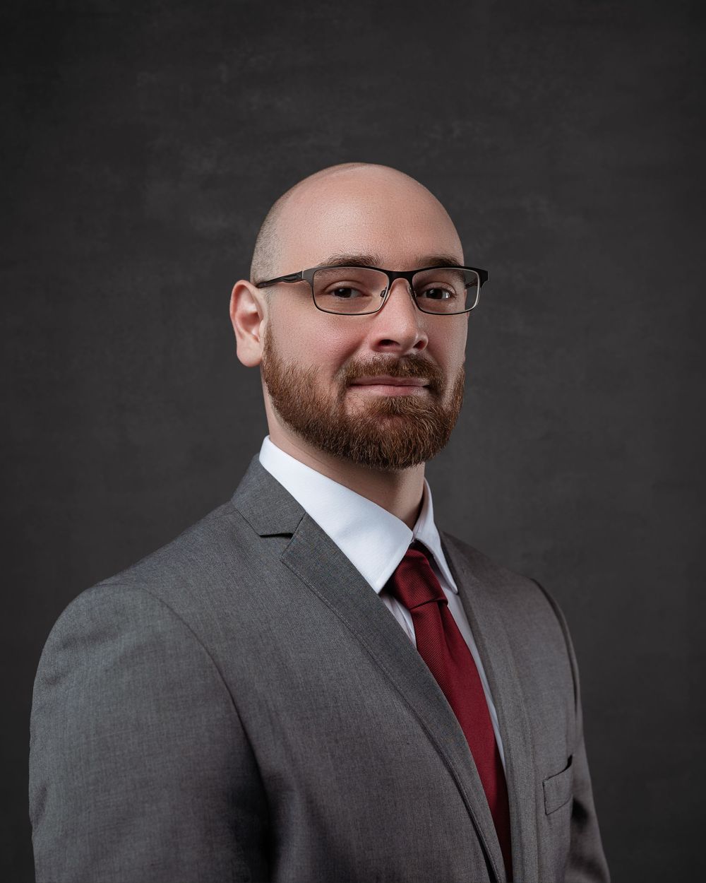 Professional headshot portrait showing a person in glasses, a grey suit jacket and red tie against a dark background.