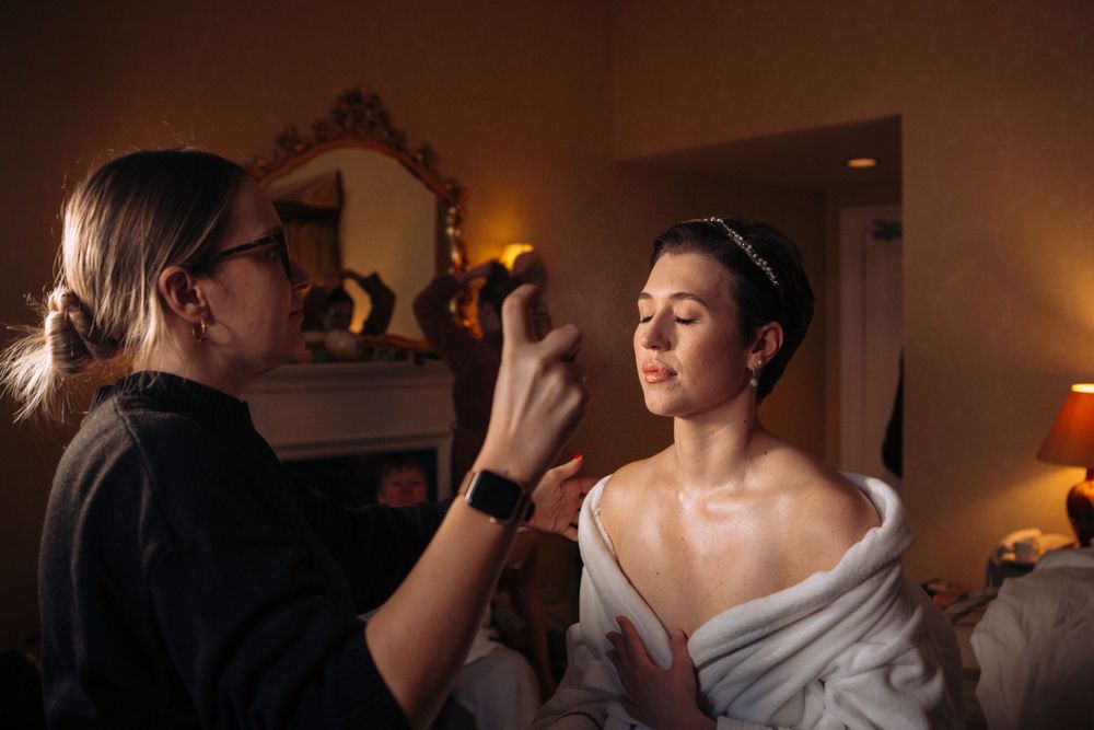 Makeup artist applying final touches during wedding preparations in warm interior lighting.