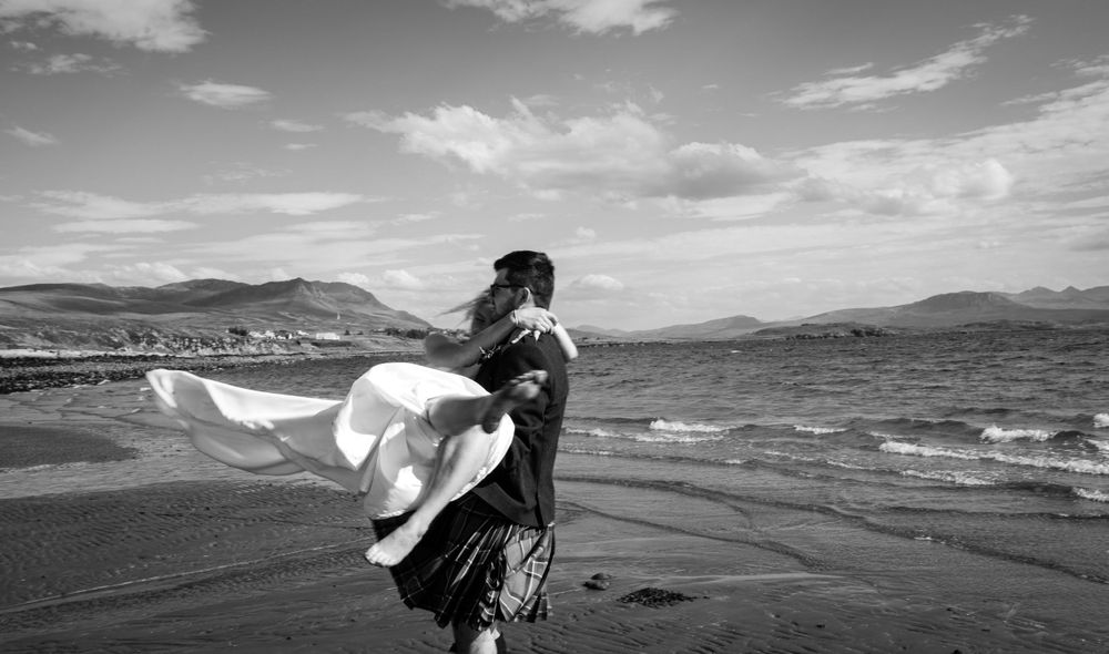 Black and white photo of couple embracing on windswept beach with mountains in distance.