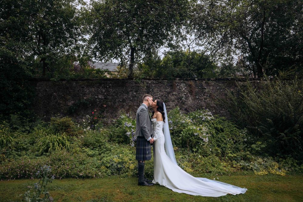 A bride and groom share a romantic kiss in a lush garden setting, with the bride wearing a flowing white gown.