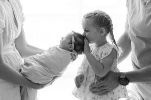 Young child gently kissing newborn sibling in white-toned family portrait.