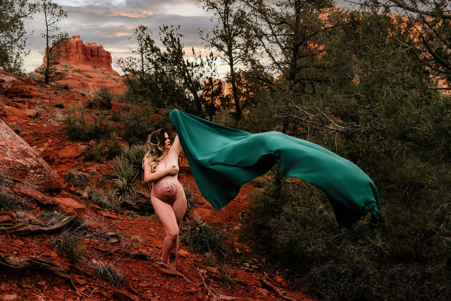 Dramatic maternity portrait at sunset with flowing teal fabric against red rock formations.