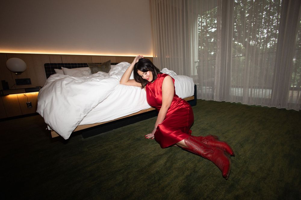 Person in flowing red dress lying on green carpet next to white bed.