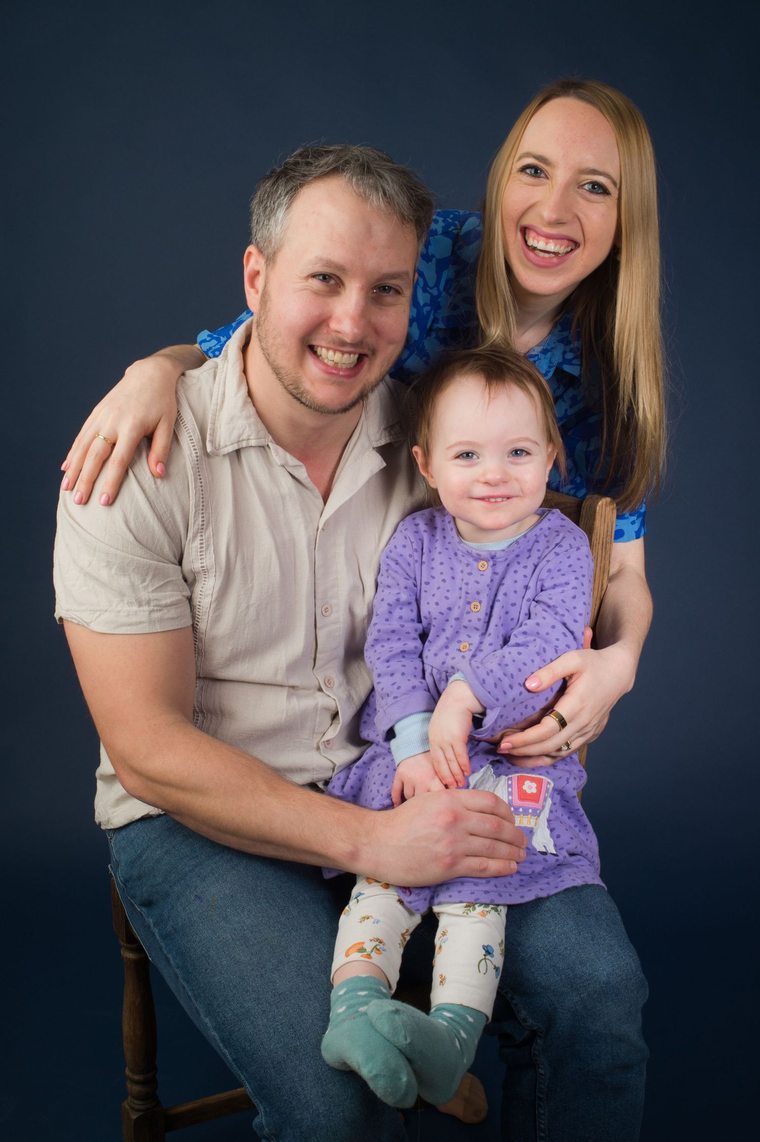 A relaxed portrait of a young family, featuring a man, woman, and baby, seated on a chair with a blue background.