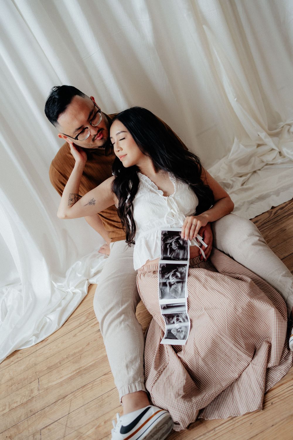 Couple sitting on floor holding ultrasound photos, sharing a tender moment during pregnancy announcement photoshoot.
