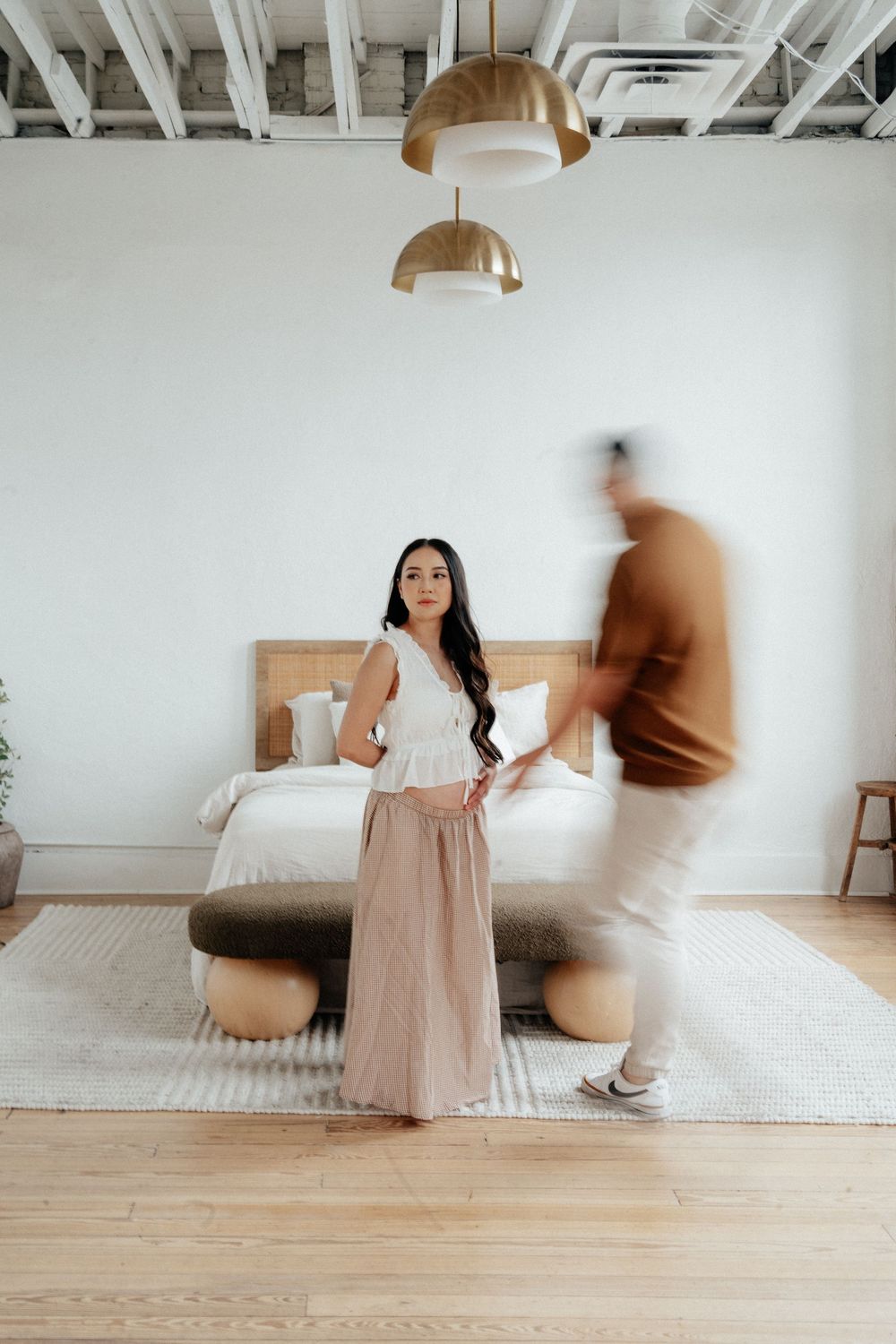 Woman in white top and pink skirt stands in modern bedroom while man in brown sweater walks past, motion blur effect.