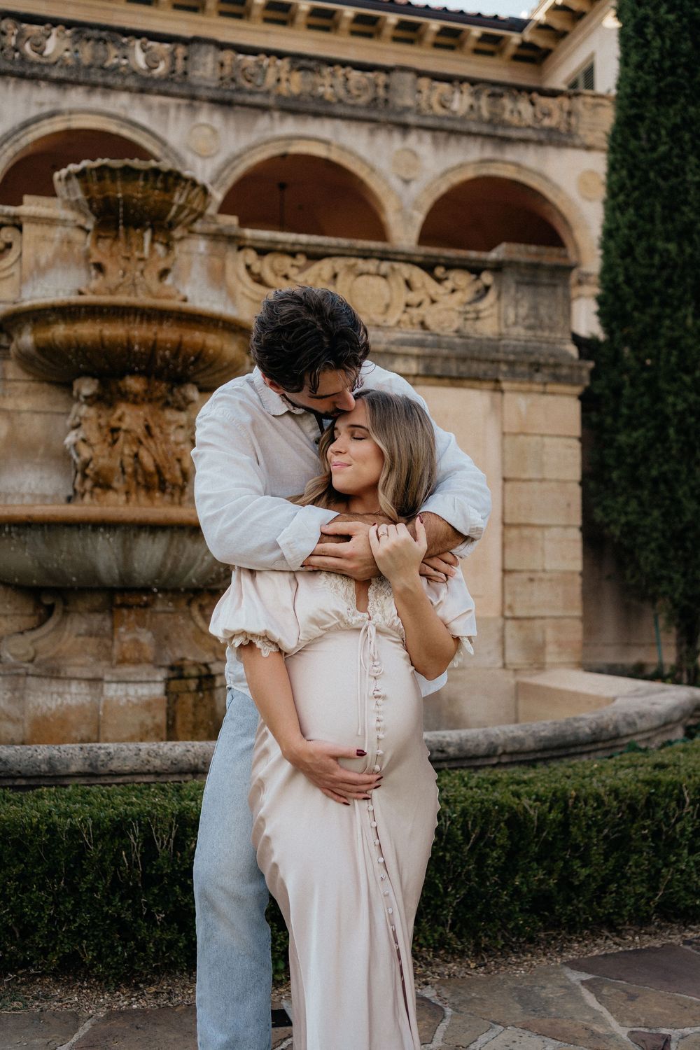Couple sharing a tender maternity portrait embrace in front of an ornate stone fountain in a garden.