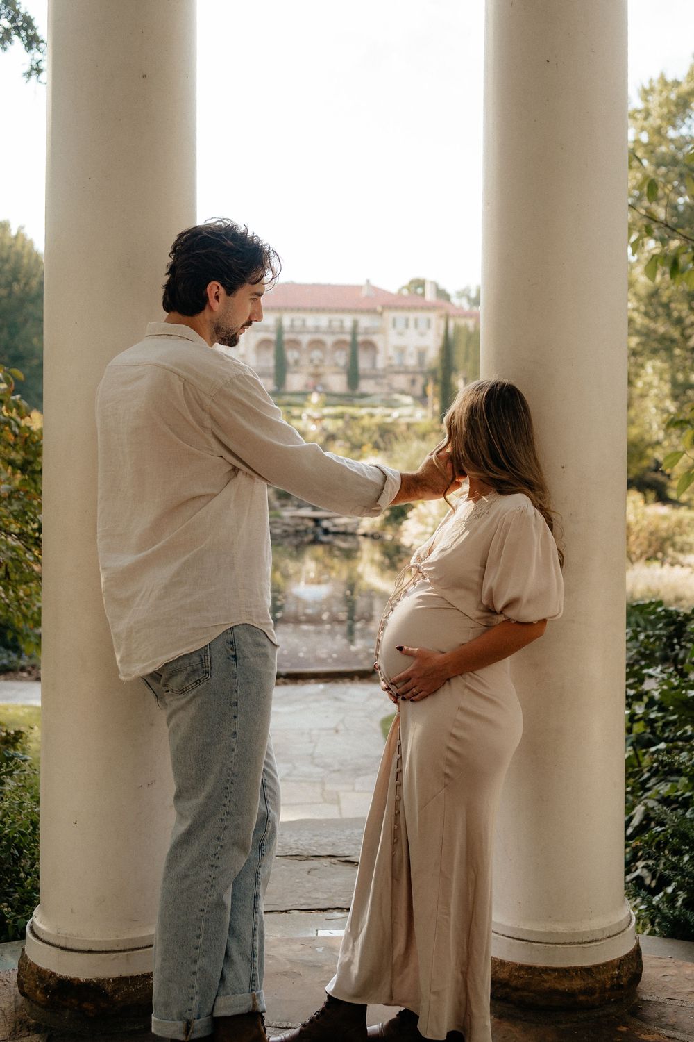 Couple sharing a tender moment between white columns in a garden, pregnant woman in beige dress cradling her baby bump.