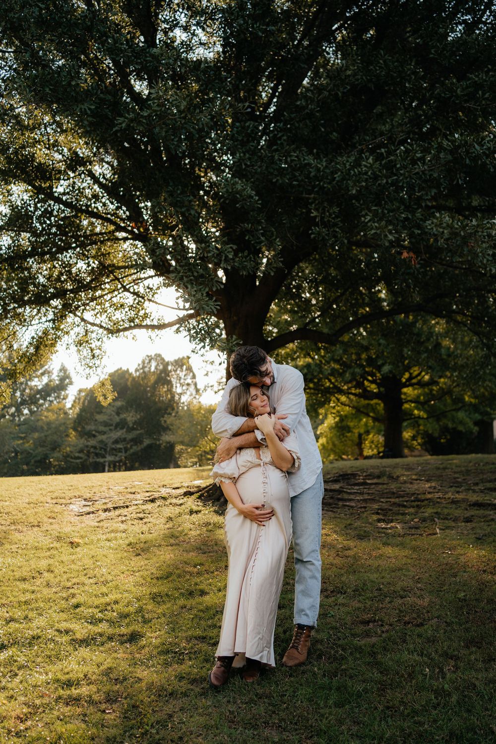 A couple embraces outdoors under a large tree at golden hour, the woman in a white dress showing a baby bump.