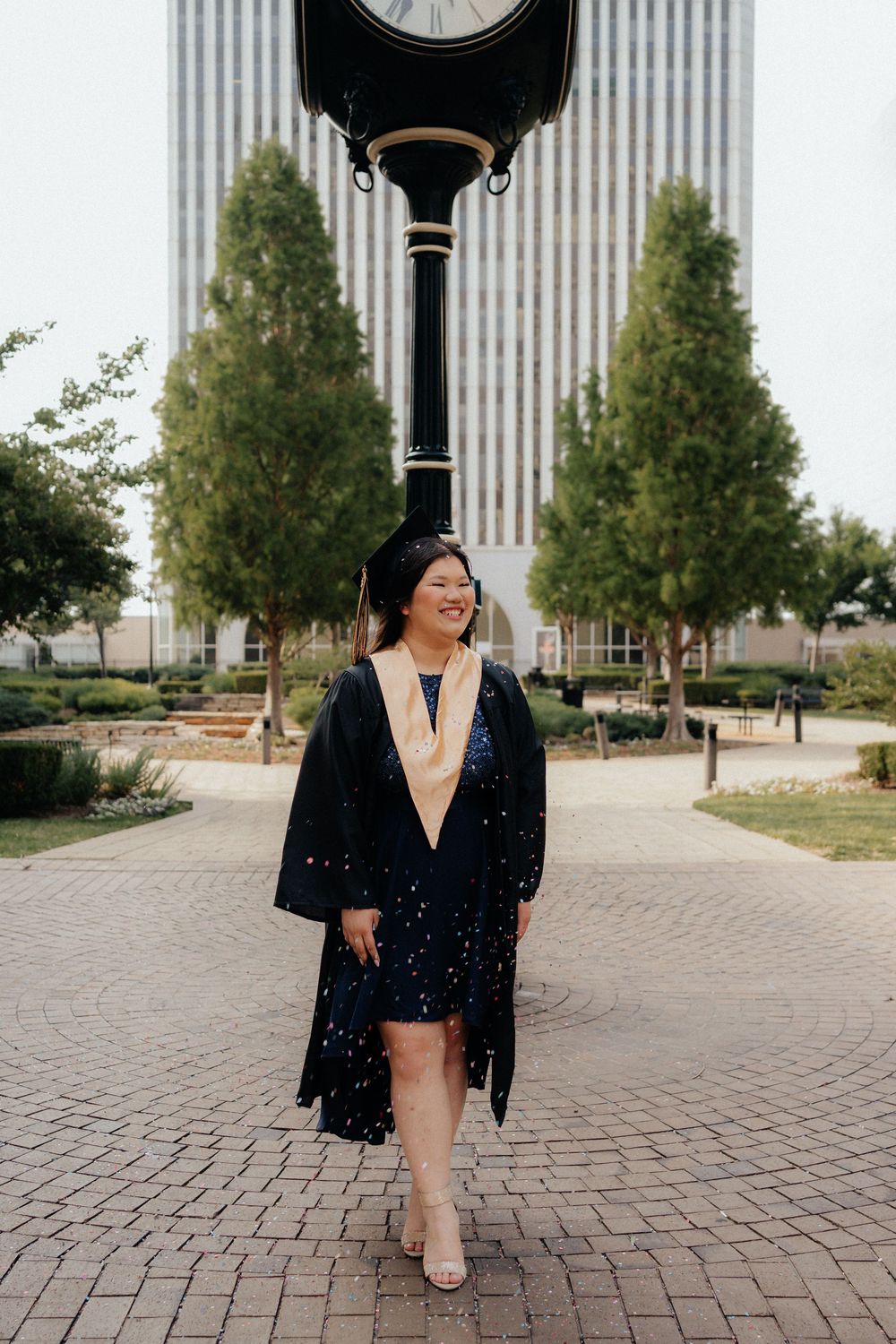Smiling graduate in black cap and gown with gold hood poses by campus clock tower on brick walkway.