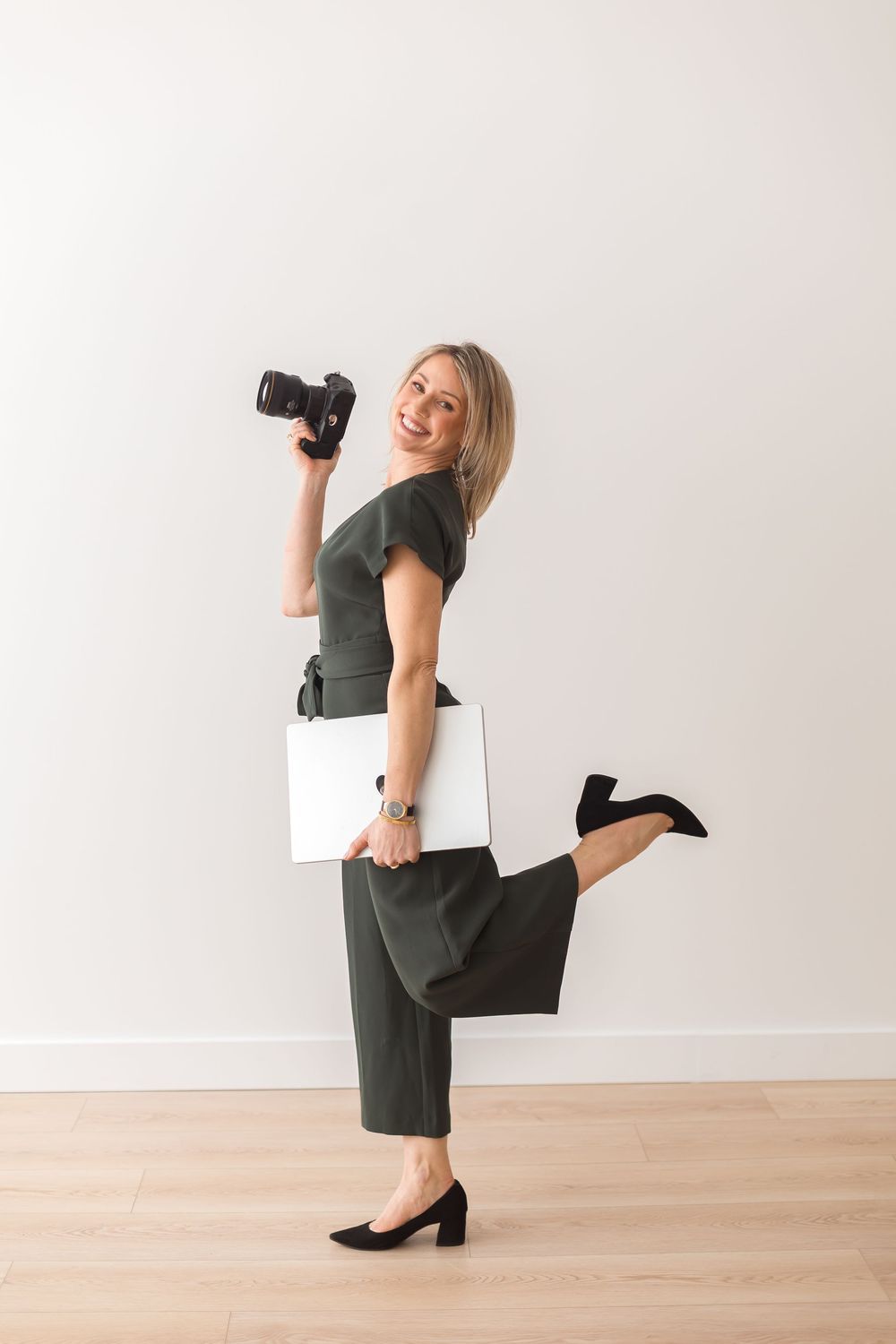 Smiling blonde woman in green jumpsuit holding DSLR camera and laptop, posing playfully on one leg against white wall.