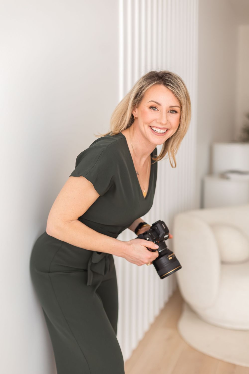 Smiling blonde woman in olive green jumpsuit holding a DSLR camera, leaning against a white wall in a bright modern studio.