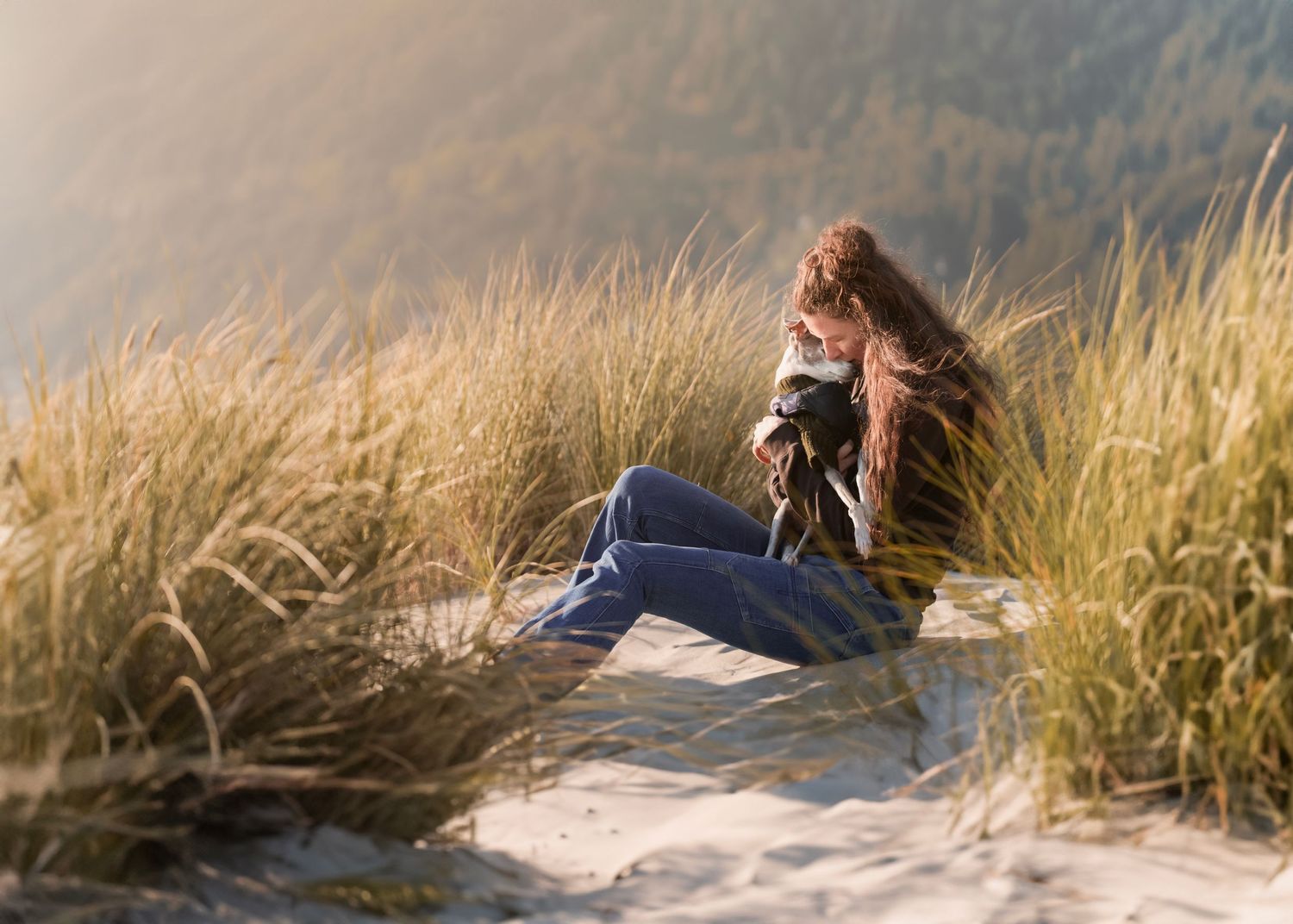 woman with the dog sitting at the sunset on the beach, dunes, grass, sunset light, hugs, long hair