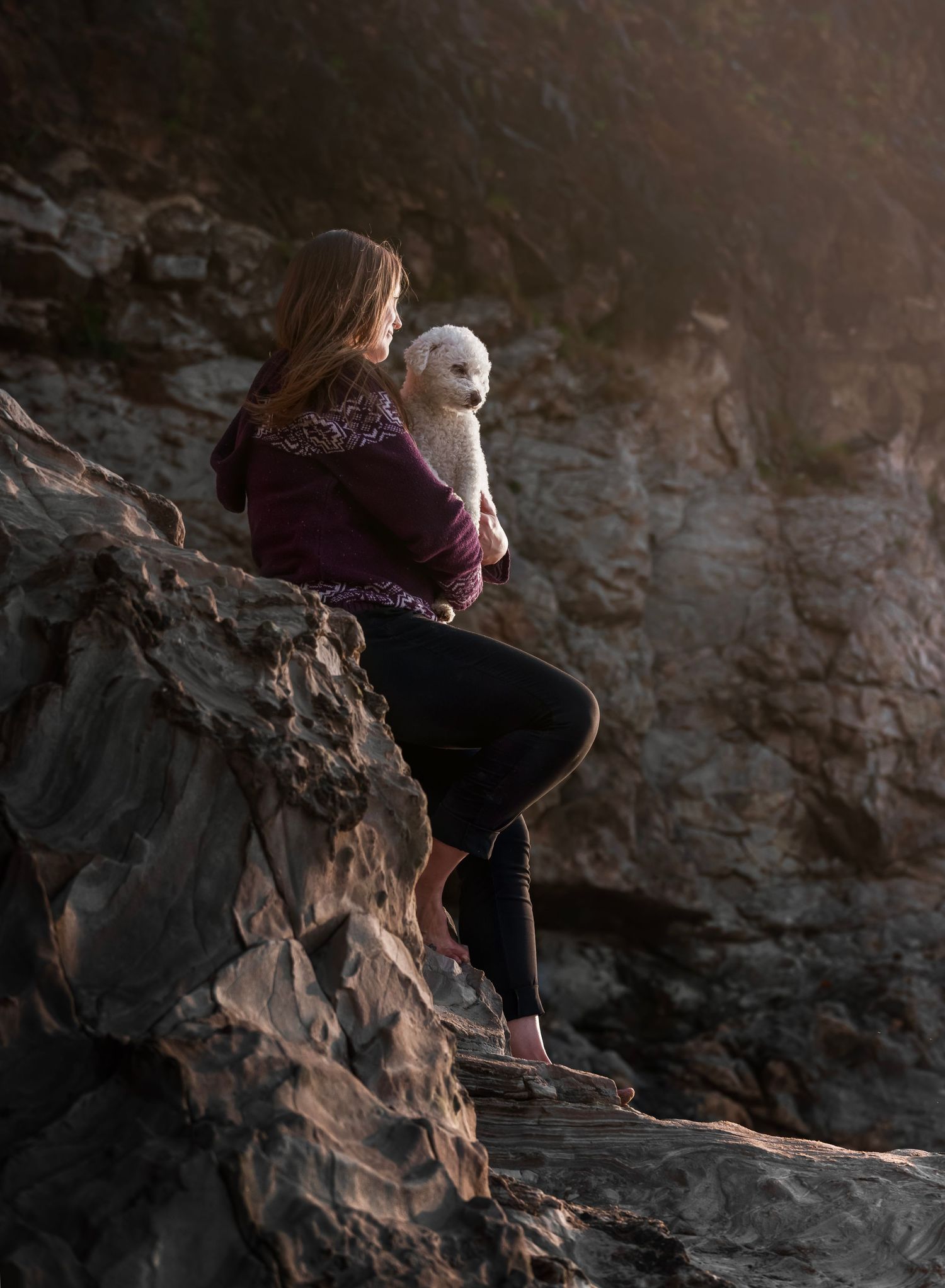 woman portrait with white small poodle dog, coast, cliffs, rocks, sunset, girl holding puppy sitting at the beach, ocean
