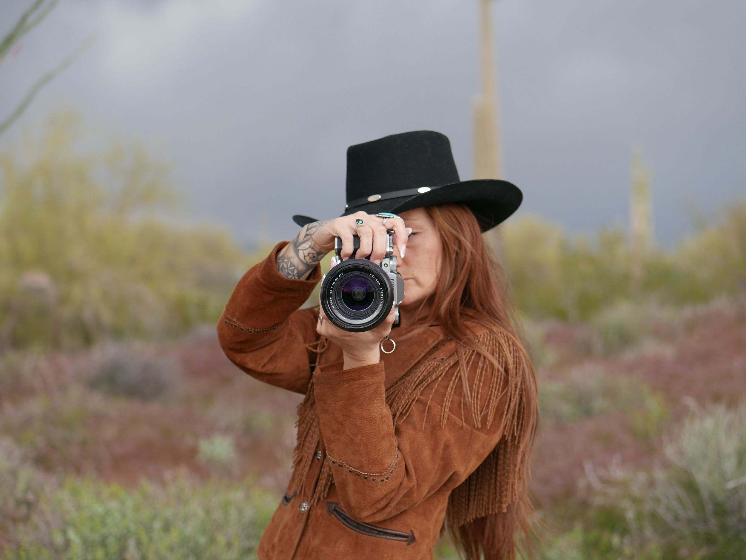 Photographer wearing brown fringe jacket and black hat captures desert scenery.