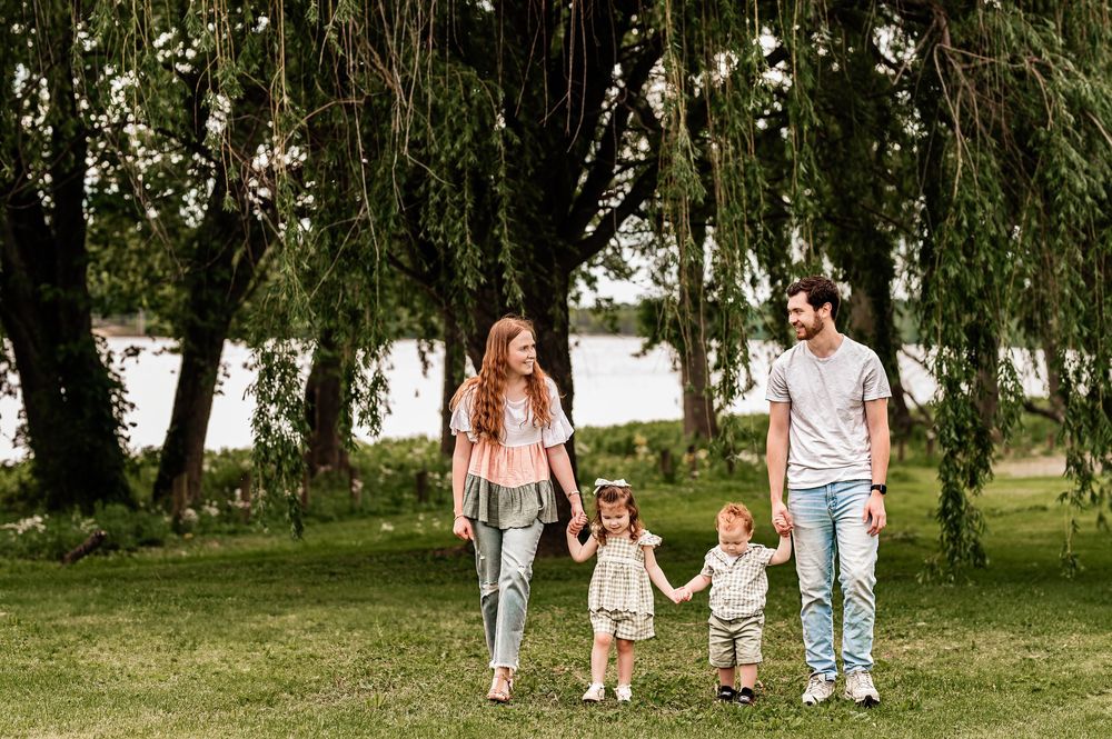 Family of four walking together under weeping willow trees by a lake.