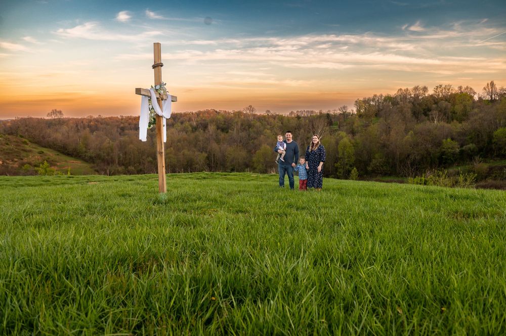Family stands silhouetted near wooden cross on hilltop during golden sunset.