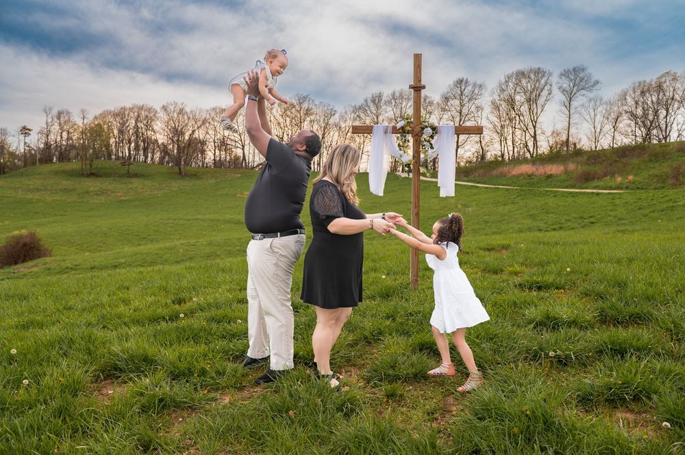 Family playing and lifting child in air on grassy hillside with crosses in background.