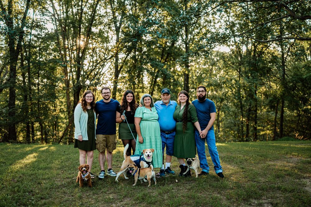 A group of people gather for outdoor family portraits in a sunlit wooded area wearing coordinated blue and teal outfits.