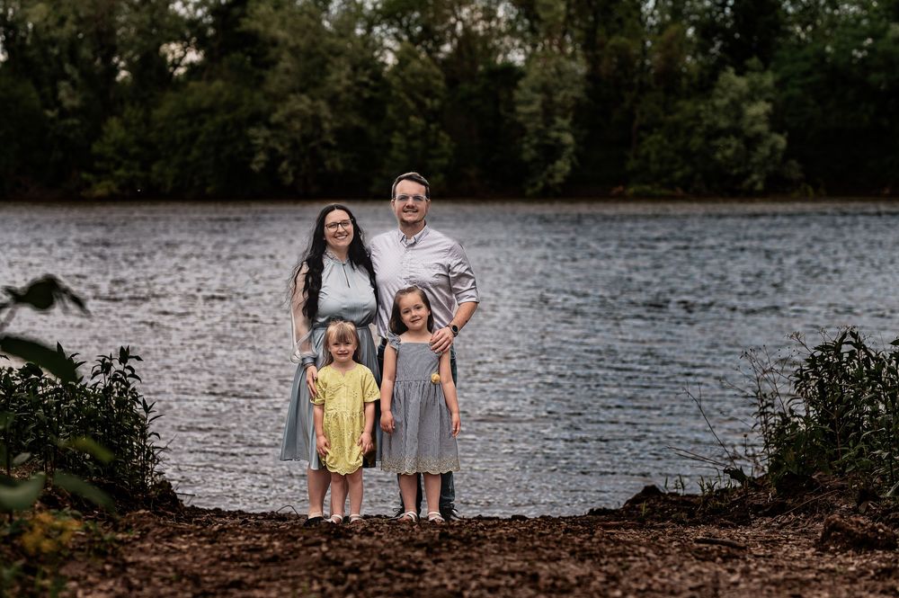 Family of four standing together near lake edge with trees in background.
