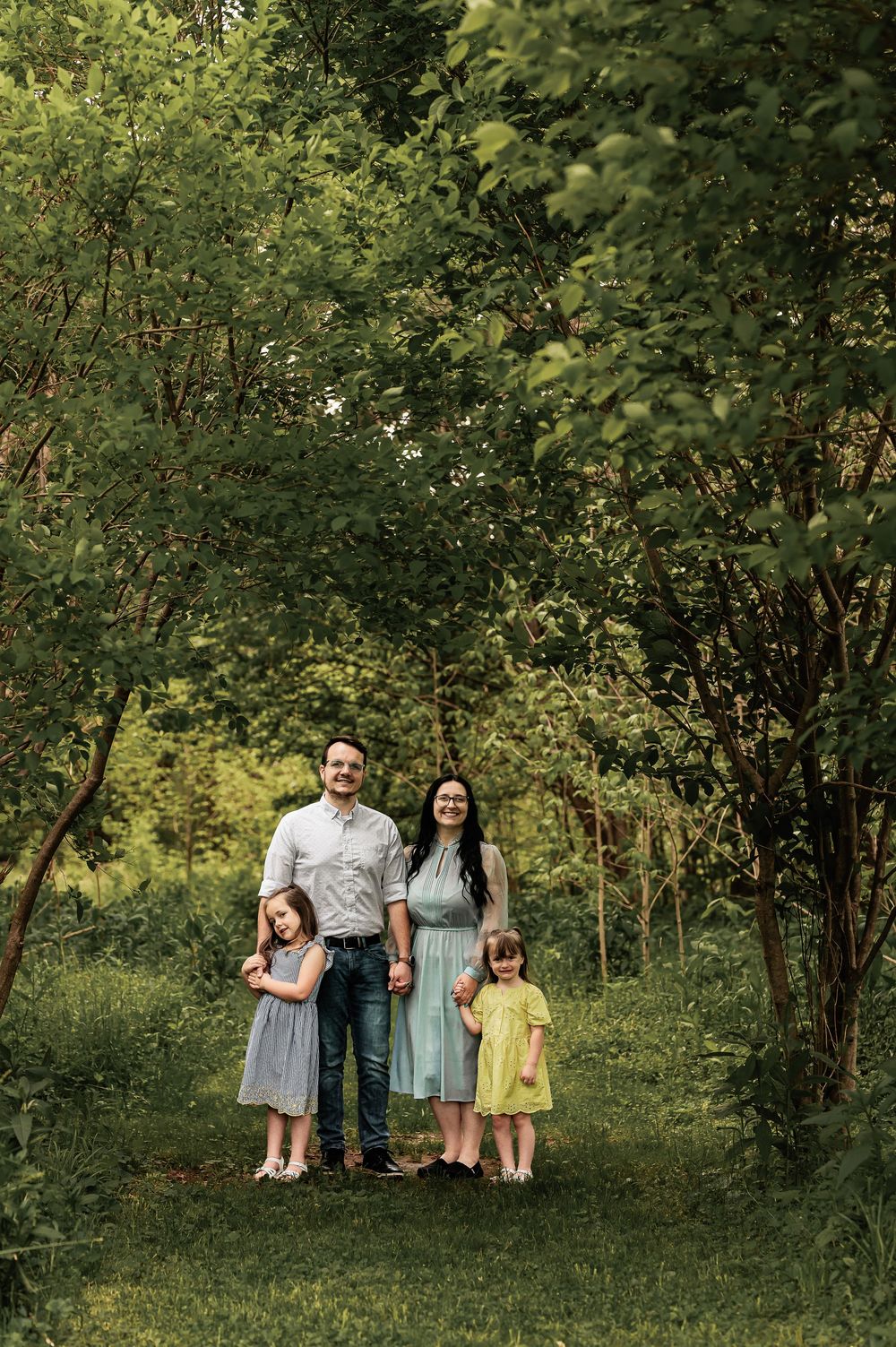 Family of four standing together on a grassy path surrounded by lush green trees.